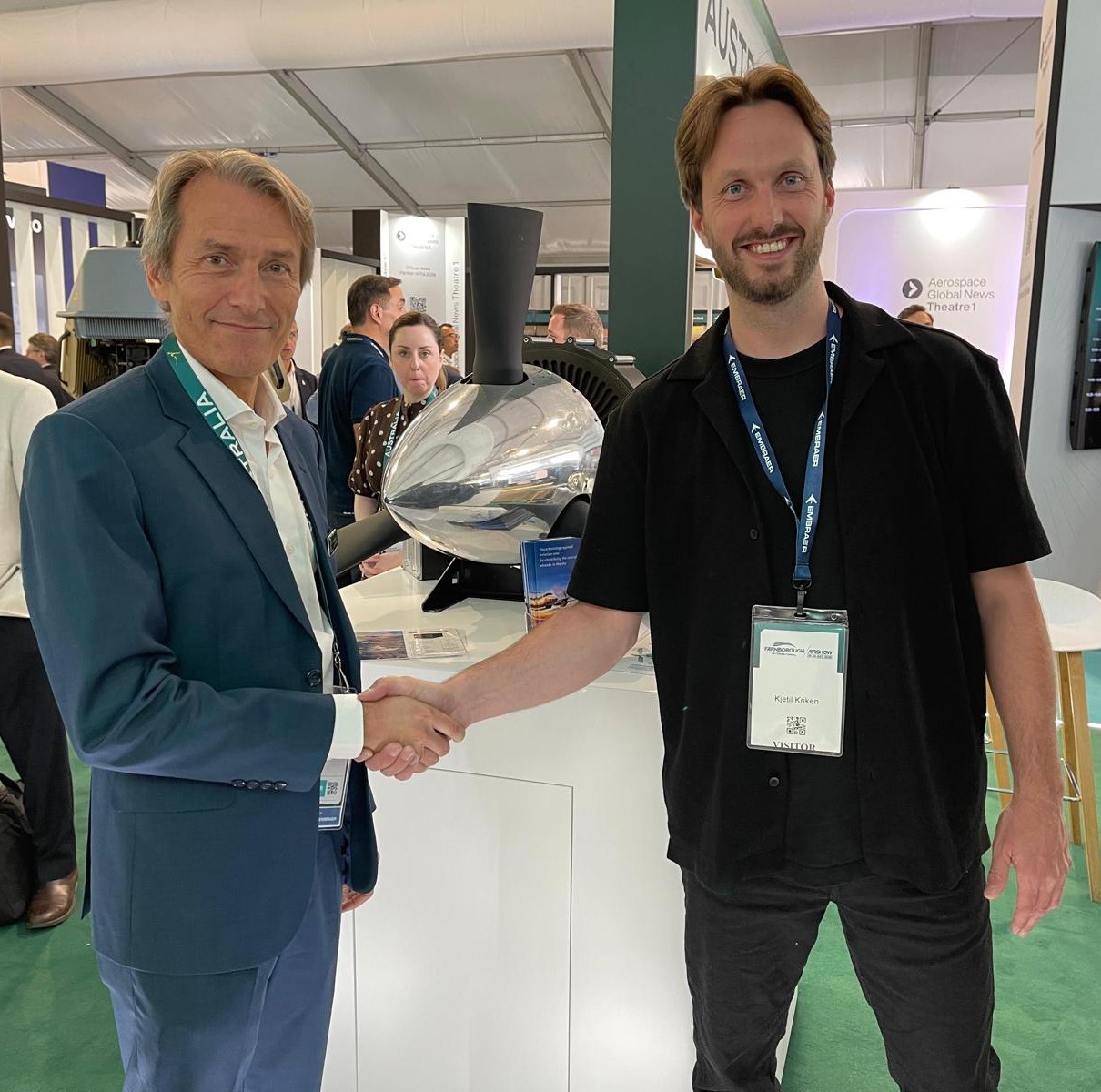 Two men shake hands at a trade show, smiling, with badges and a large machine in the background.