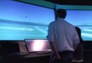 Wisk and NASA Simulate Multi-Aircraft Autonomous Flight Operations in Controlled Airspace Two people stand at a flight-simulation setup, facing a large curved screen showing a blue sky, runway, and a small airplane in the distance.