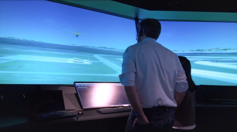 Wisk and NASA Simulate Multi-Aircraft Autonomous Flight Operations in Controlled Airspace Two people stand at a flight-simulation setup, facing a large curved screen showing a blue sky, runway, and a small airplane in the distance.