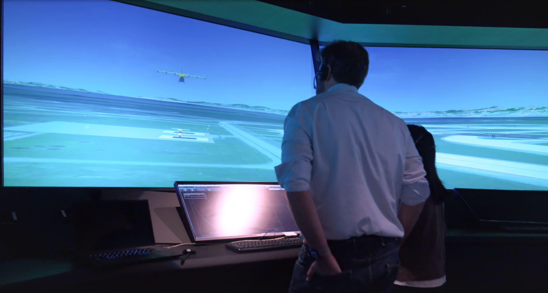 Two people stand at a flight-simulation setup, facing a large curved screen showing a blue sky, runway, and a small airplane in the distance.