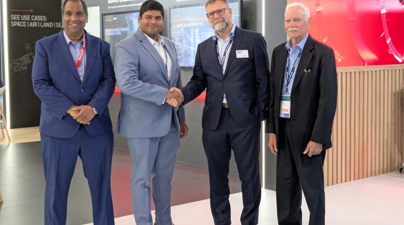 Four men in suits posing and shaking hands at a trade-show booth in a modern display area.