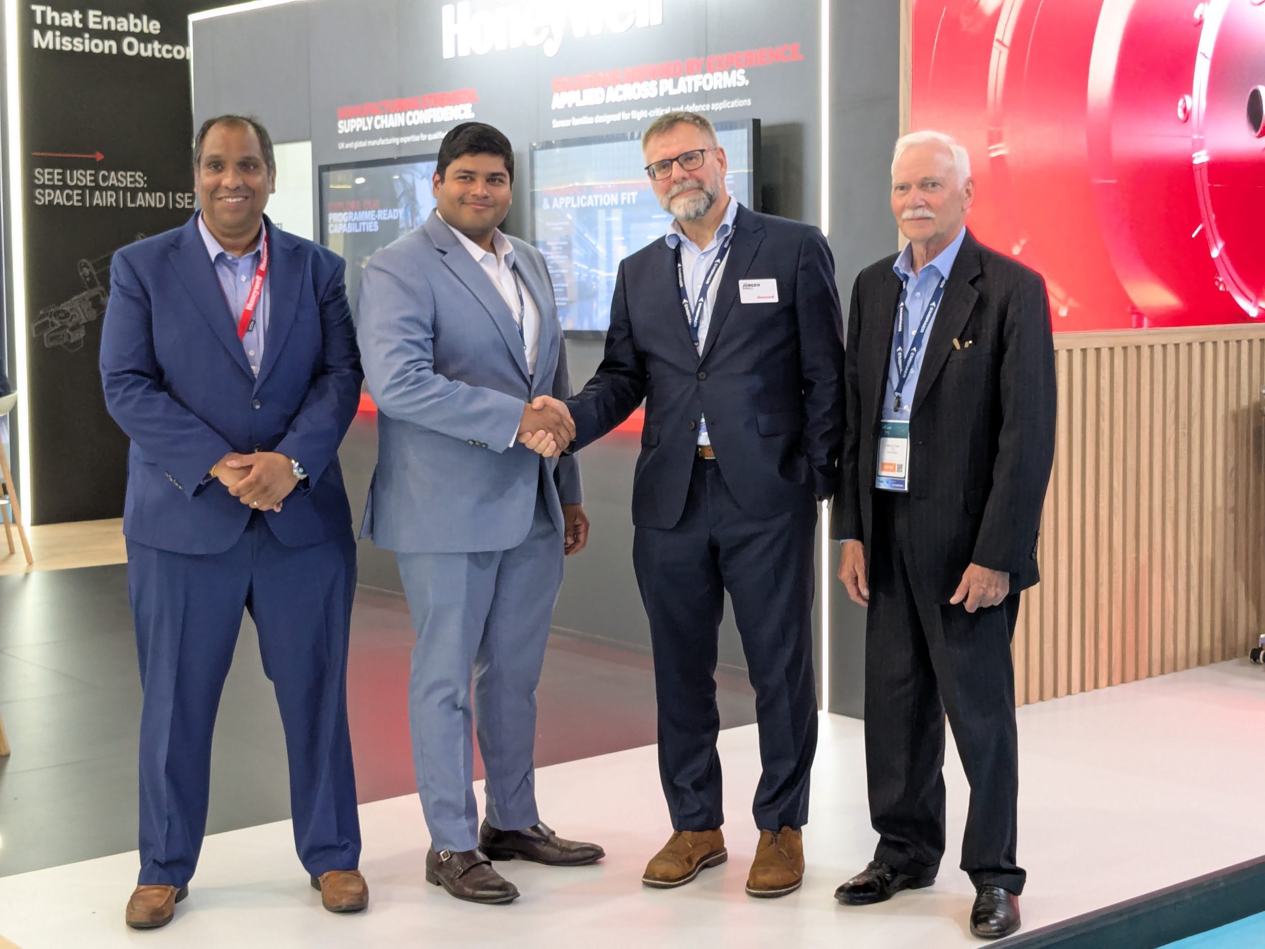 Four men in suits posing and shaking hands at a trade-show booth in a modern display area.
