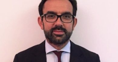 Professional headshot: man with glasses, dark suit, white shirt, maroon tie against a pale background.