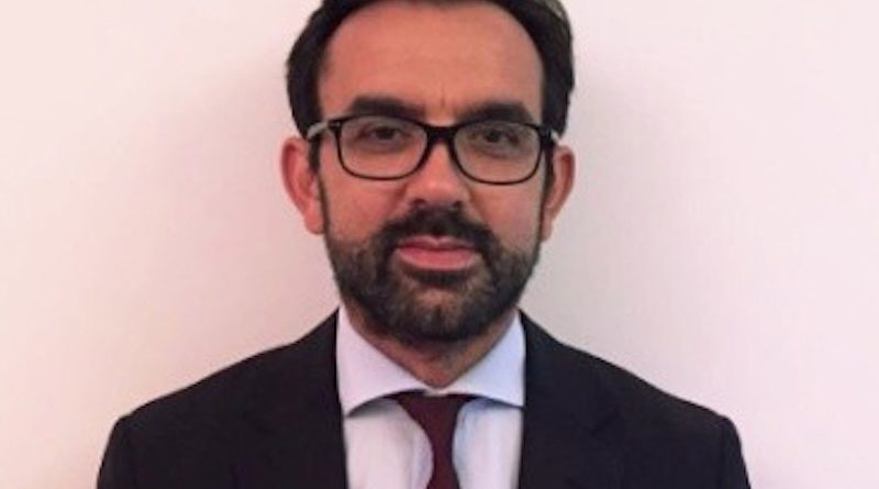 Professional headshot: man with glasses, dark suit, white shirt, maroon tie against a pale background.