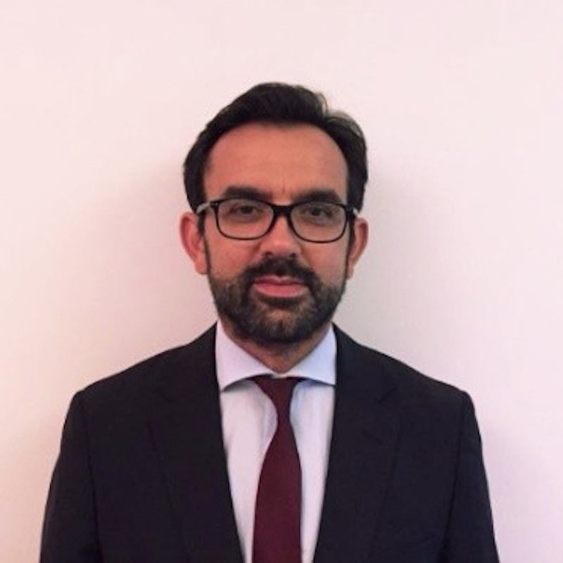Professional headshot: man with glasses, dark suit, white shirt, maroon tie against a pale background.