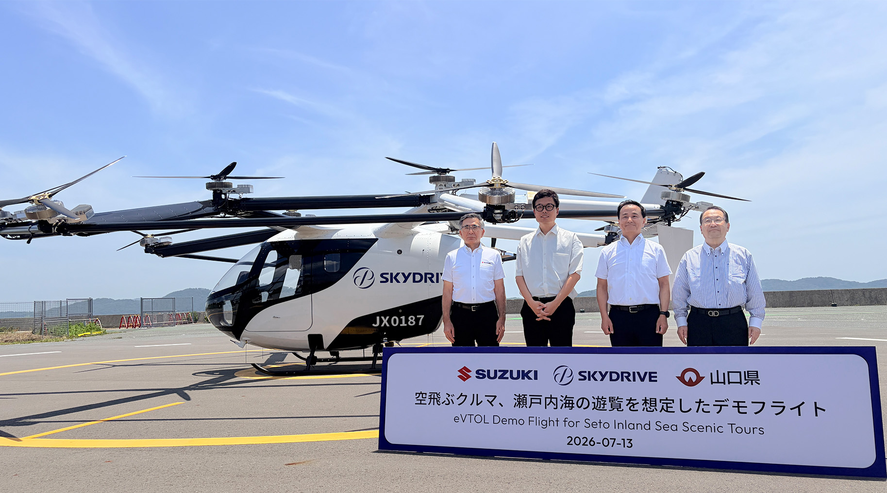 Four men in white shirts stand beside a SkyDrive eVTOL helicopter on a runway, with a promotional sign in front about Seto Inland Sea tours (dated 2026-07-13).