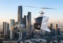 White autonomous rotorcraft hovering above a dense city skyline at sunset.