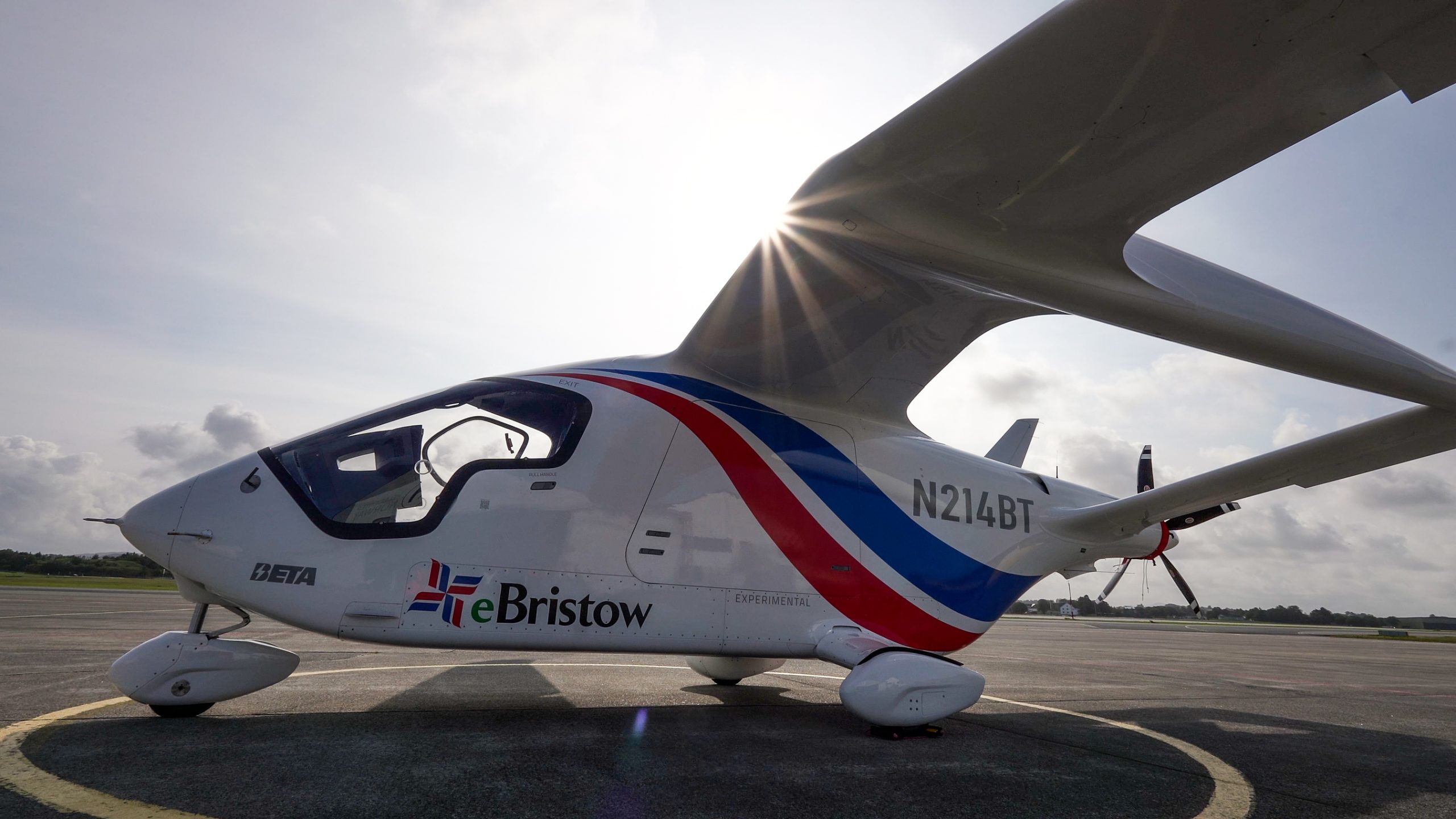 Small white light aircraft on a tarmac with red–blue stripes and sun bursting behind the wing behind the cockpit window area.