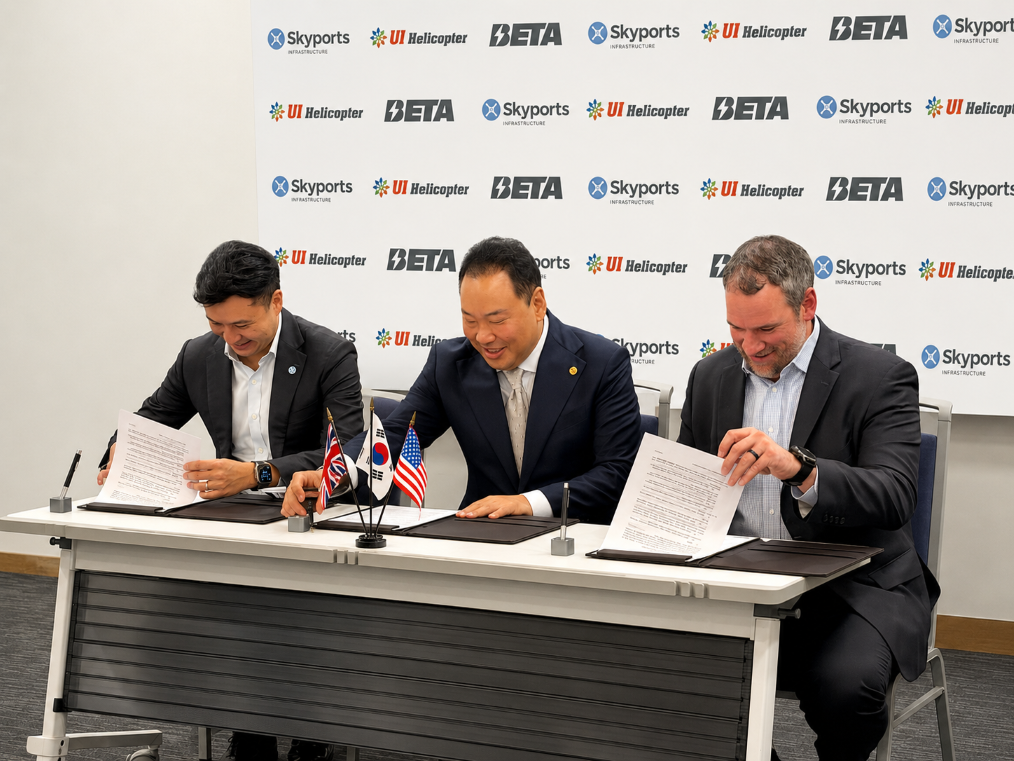 Three businessmen in suits sign documents at a table, with UK, South Korea, and US flags in front and a branded backdrop behind them.