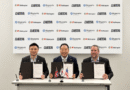 Three men in suits sit at a table holding signed documents during a formal agreement ceremony, flags in front, backdrop with logos behind them.