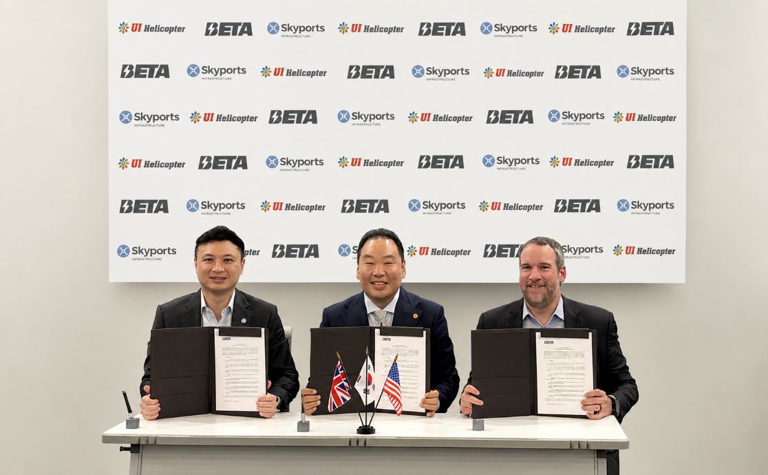 Three men in suits sit at a table holding signed documents during a formal agreement ceremony, flags in front, backdrop with logos behind them.