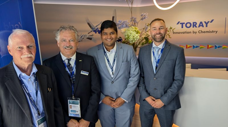 Four men in suits with lanyards posing at a Toray display booth with a floral backdrop and waving banner accents in the background.