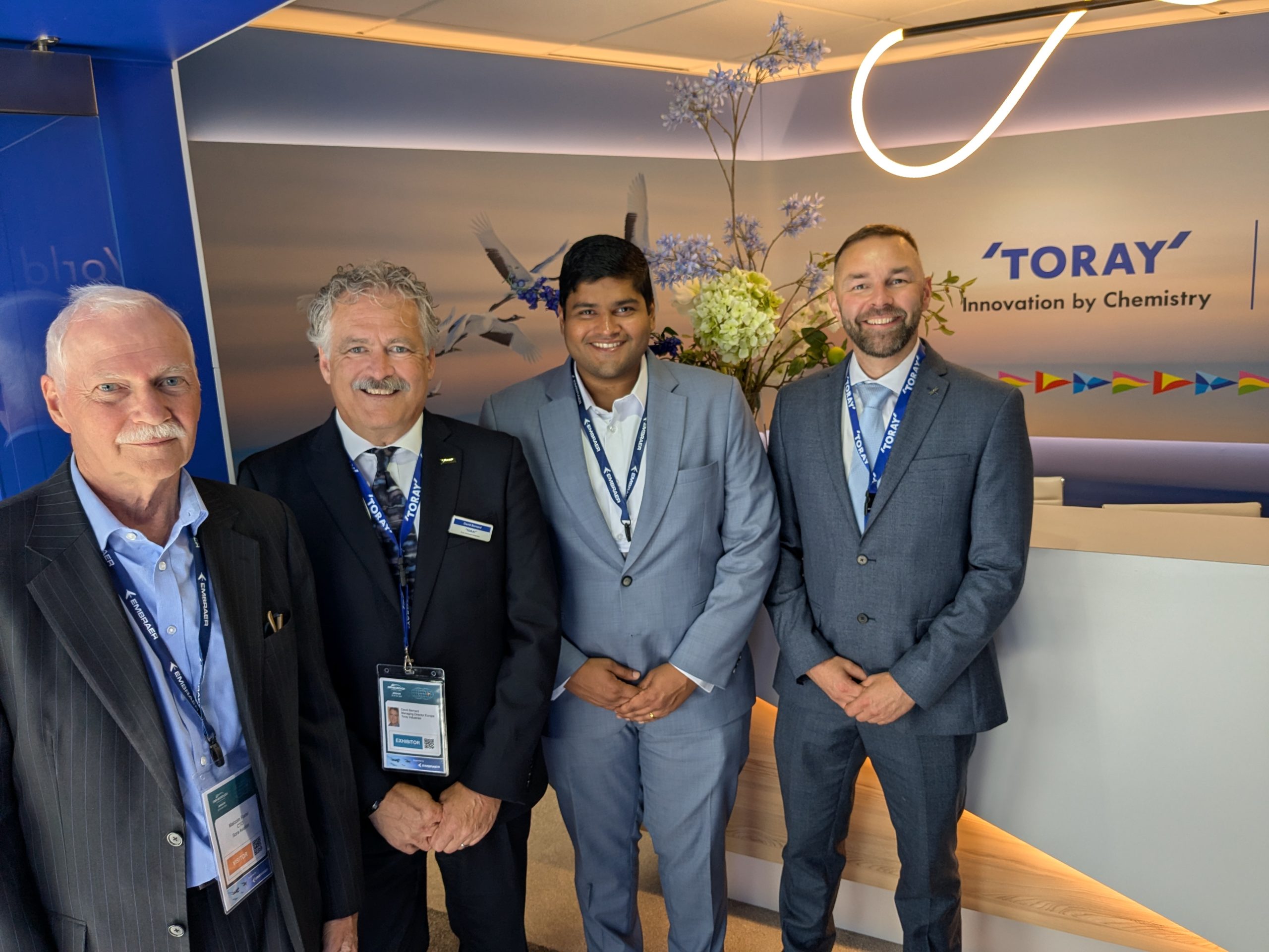 Four men in suits with lanyards posing at a Toray display booth with a floral backdrop and waving banner accents in the background.