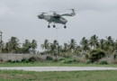 Sylla Flight Test Campaign - eVTOL Insights Large four-rotor drone hover-lift with a tether descending to the ground over a concrete pad, palm trees in the background.