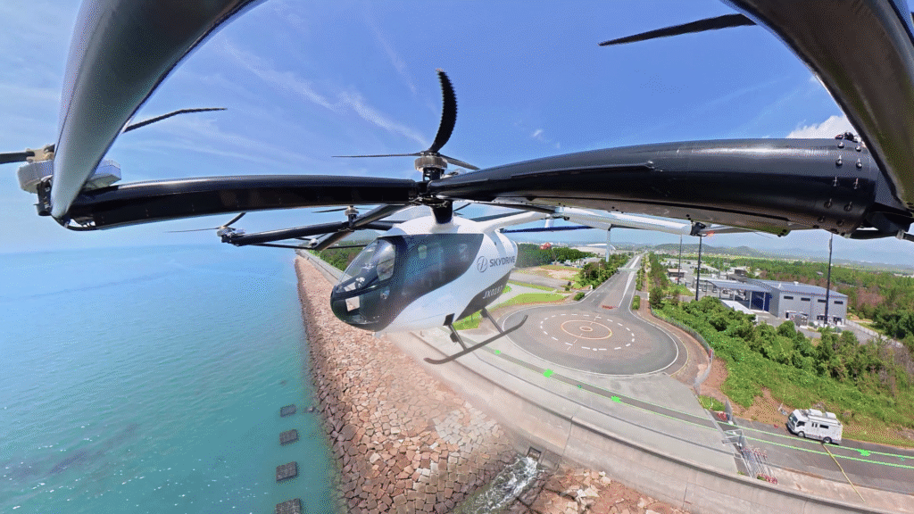 Thumbnail of the video - eVTOL Insights Aerial view from a small helicopter over a coast, showing the rotor hub and edge of the windscreen with a seawall to the left and a road with buildings to the right.