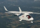White small aircraft in flight over a patchwork countryside, two people visible in the cockpit smiling at the camera, registration N336MR.