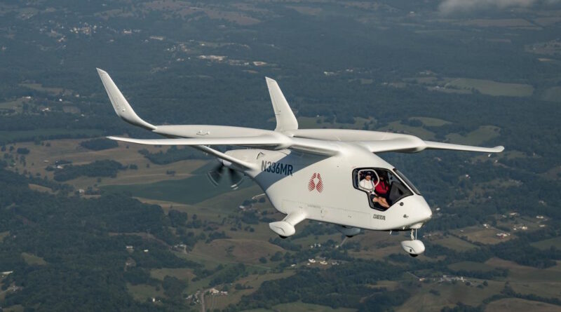 White small aircraft in flight over a patchwork countryside, two people visible in the cockpit smiling at the camera, registration N336MR.