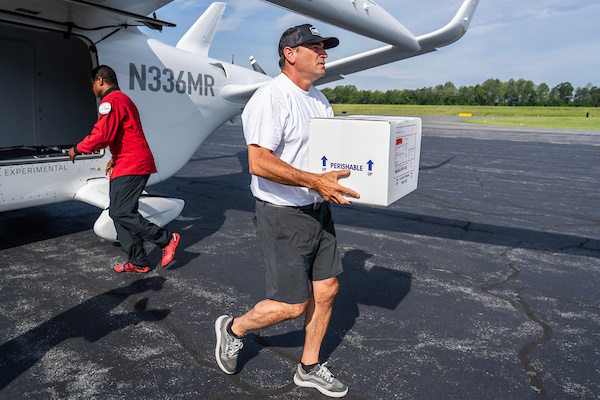 Man in a cap carries a white box labeled Perishable toward a small airplane on a tarmac; another person in red walks toward the aircraft.
