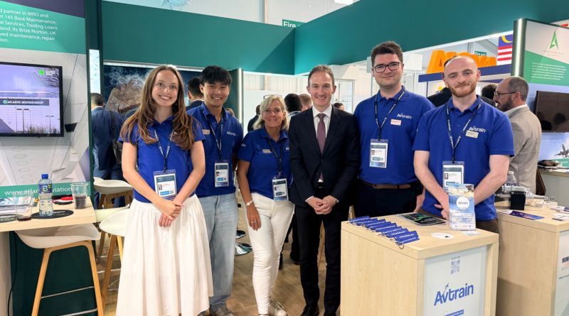 avtrain-farnborough - eVTOL Insights Group of staff at a trade show booth posing for a photo; several wearing blue Avtrain polo shirts with lanyards and badges.