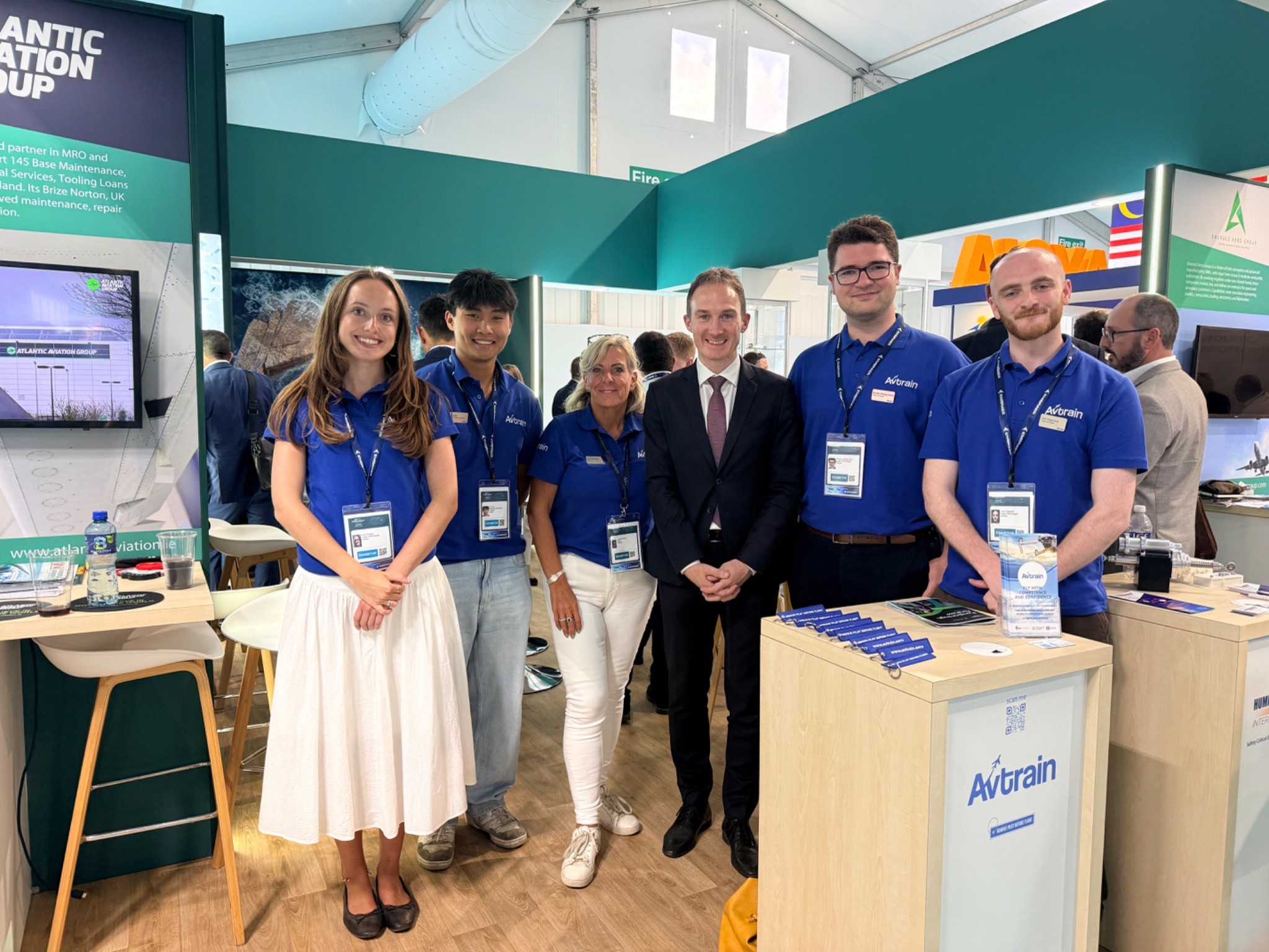 Group of staff at a trade show booth posing for a photo; several wearing blue Avtrain polo shirts with lanyards and badges.