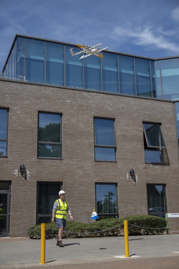 Construction worker in a yellow safety vest and hard hat walking on the sidewalk outside a brick building; a white drone hovers above the glass-roof extension.