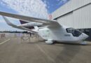 e-Smart BETA ALIA CTOL 1 - eVTOL Insights White small aircraft with N214BT registration parked on tarmac beside a corrugated metal hangar, e-Smart Avia branding visible on fuselage