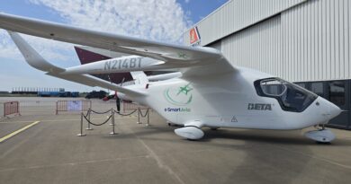 White small aircraft with N214BT registration parked on tarmac beside a corrugated metal hangar, e-Smart Avia branding visible on fuselage