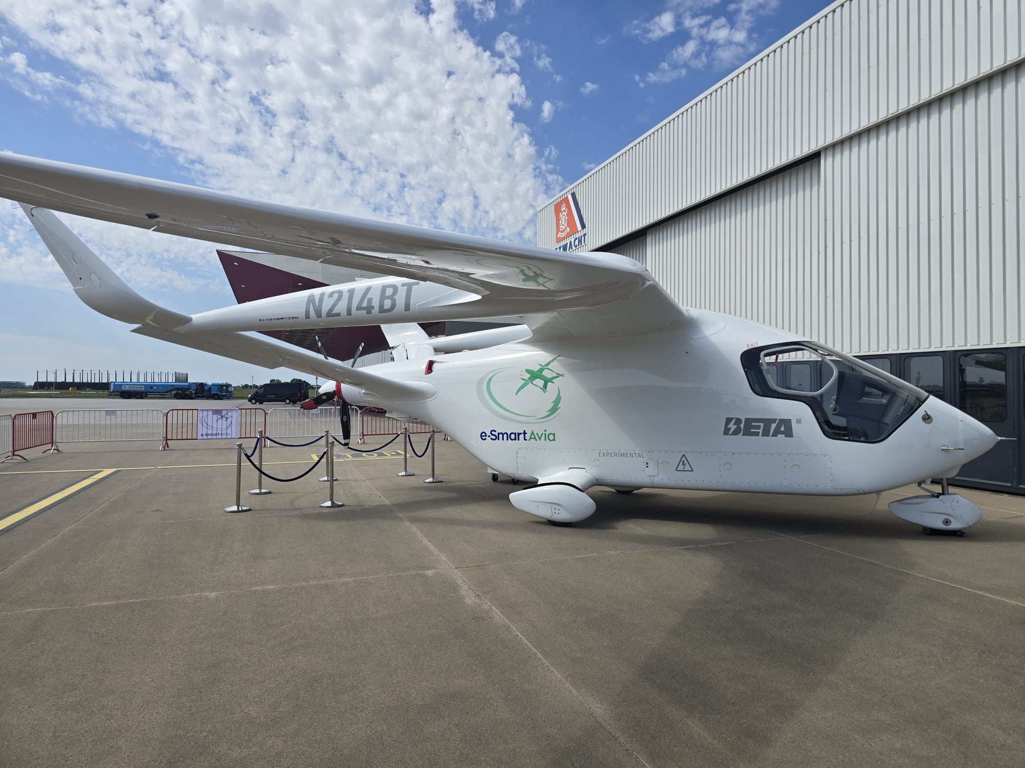 White small aircraft with N214BT registration parked on tarmac beside a corrugated metal hangar, e-Smart Avia branding visible on fuselage