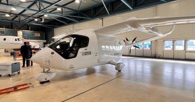 Interior of a large aircraft hangar with a white, futuristic plane in the foreground and two men in suits inspecting it, a second plane visible in the background