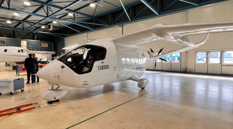 Interior of a large aircraft hangar with a white, futuristic plane in the foreground and two men in suits inspecting it, a second plane visible in the background