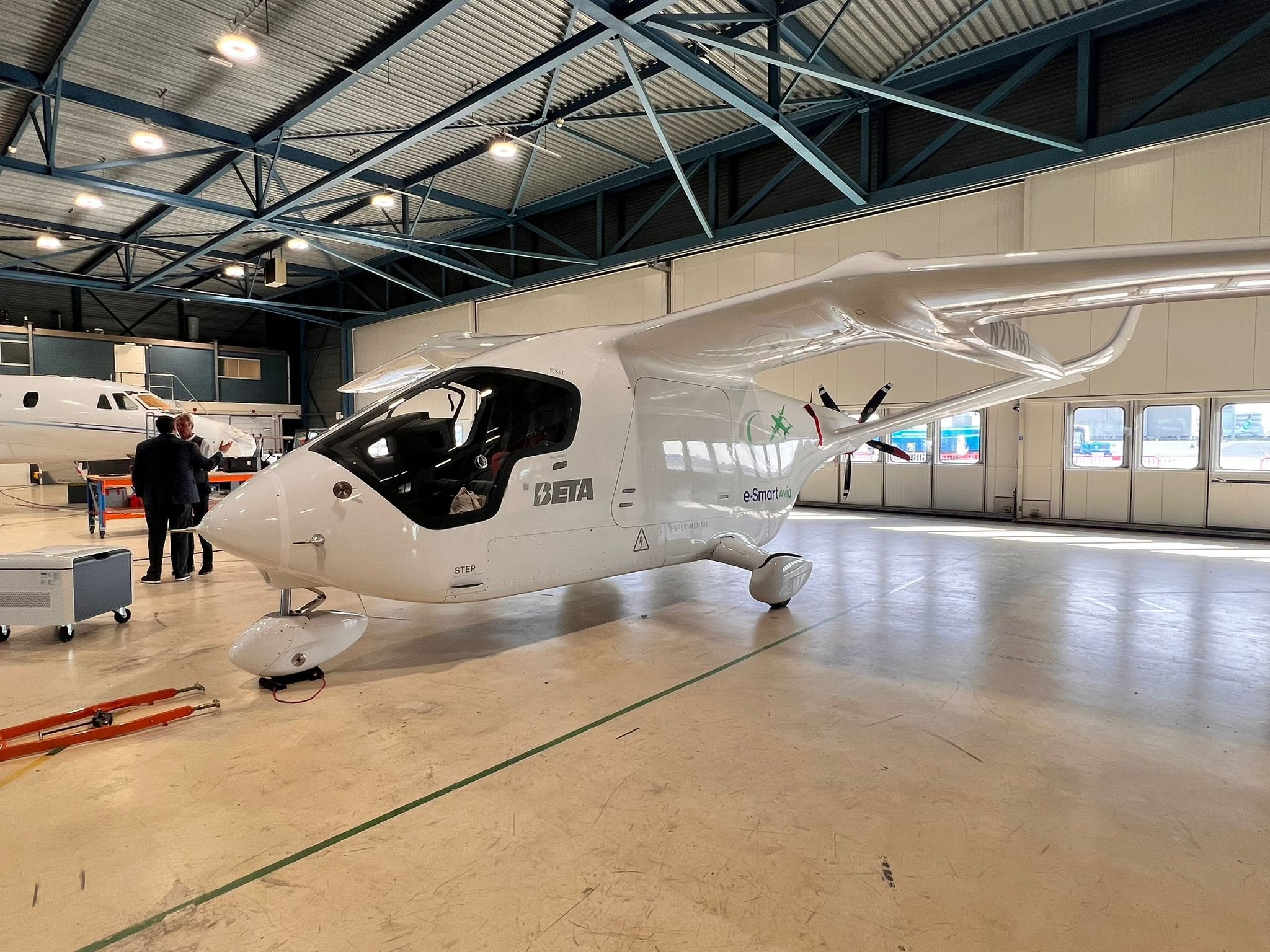 Interior of a large aircraft hangar with a white, futuristic plane in the foreground and two men in suits inspecting it, a second plane visible in the background