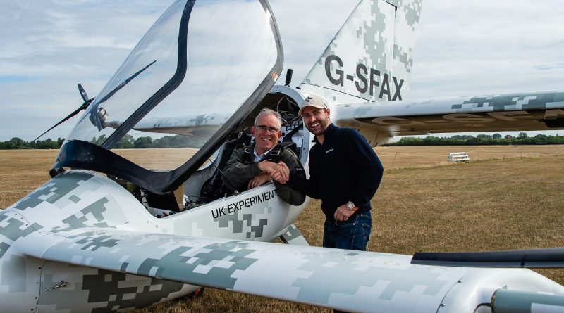 Two men pose beside a camouflaged white experimental airplane with the cockpit open on a grass airfield, tail numbered G-SFAX.
