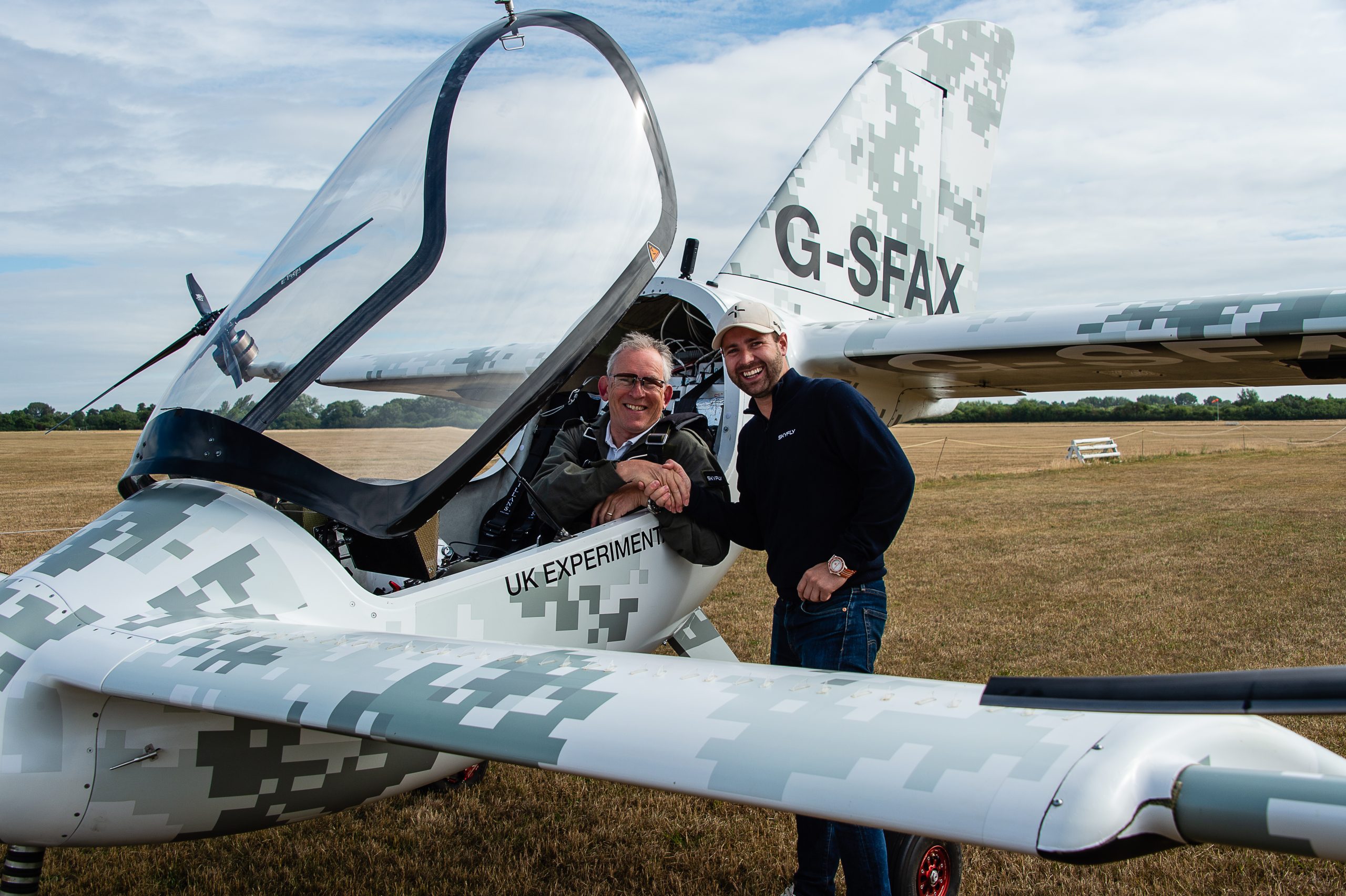 Two men pose beside a camouflaged white experimental airplane with the cockpit open on a grass airfield, tail numbered G-SFAX.