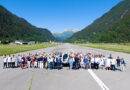 Group of people posed on a runway with a white helicopter in the center, mountains and blue sky in the background.