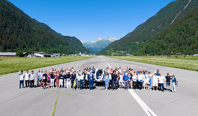 Group of people posed on a runway with a white helicopter in the center, mountains and blue sky in the background.