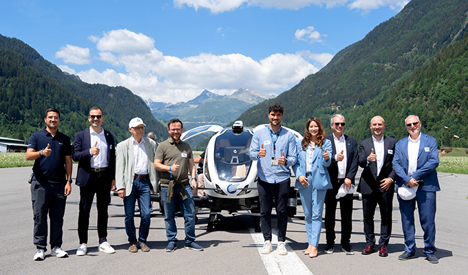 Group of professionals standing in front of a small airplane on a runway, giving thumbs up with mountains in the background, sunny day.