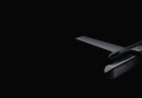 Dark, minimal photo of a sleek drone visible on the right against a black background, showing its wings and propellers.