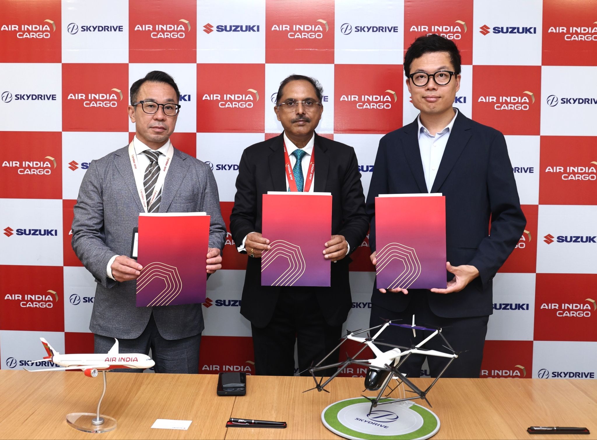 Three men in suits hold red folders at a formal event, standing before a red-and-white branded backdrop (Air India Cargo, Suzuki).