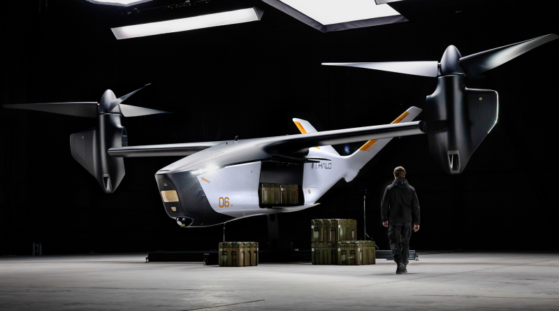 Indoor display of a futuristic white aircraft with oversized rotors in a dark hangar; a person in a jumpsuit stands nearby for scale amid cargo crates.
