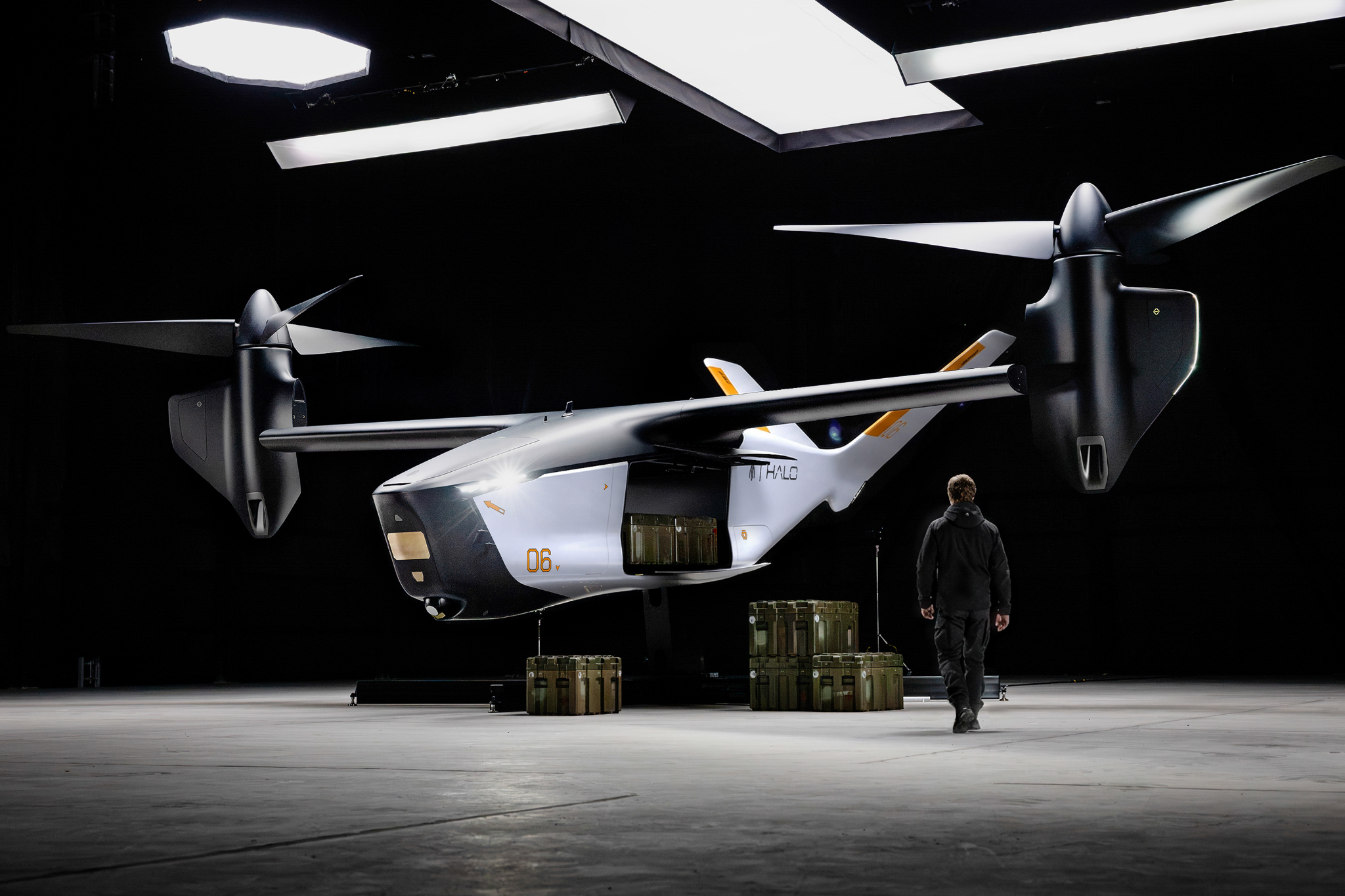 Indoor display of a futuristic white aircraft with oversized rotors in a dark hangar; a person in a jumpsuit stands nearby for scale amid cargo crates.
