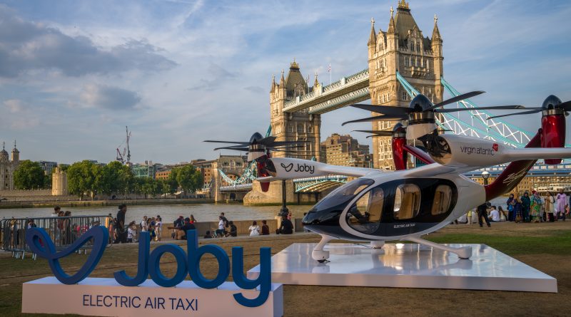 Electric air taxi helicopter on a white display platform by the River Thames with Tower Bridge in the background and visitors nearby in London.