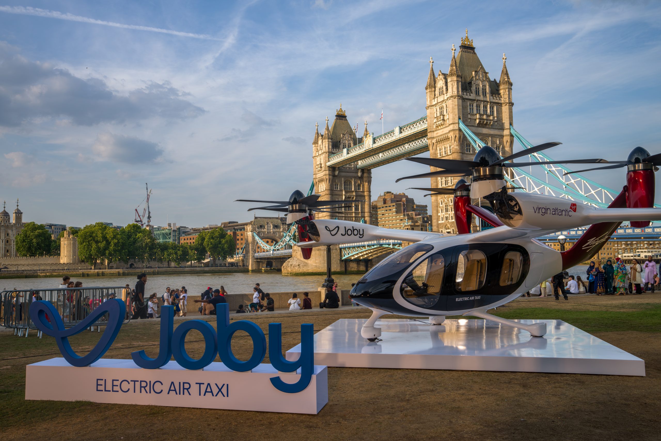 Electric air taxi helicopter on a white display platform by the River Thames with Tower Bridge in the background and visitors nearby in London.