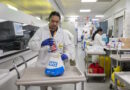 Lab technician in a white coat handling a specimen bag labeled NHS at a bench in a busy hospital laboratory with other staff in the background.