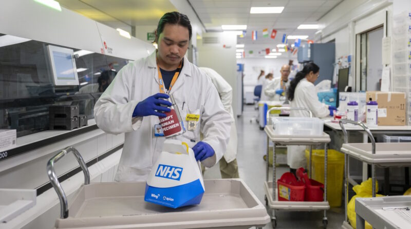 Lab technician in a white coat handling a specimen bag labeled NHS at a bench in a busy hospital laboratory with other staff in the background.