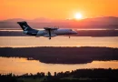 Twin-engine turboprop aircraft in landing/approach over a sunset-lit landscape with water and hills in the background.