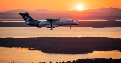 Twin-engine turboprop aircraft in landing/approach over a sunset-lit landscape with water and hills in the background.