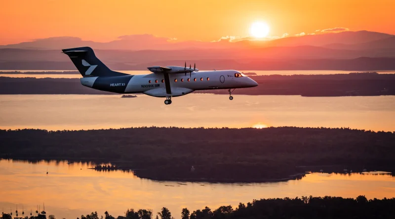 Twin-engine turboprop aircraft in landing/approach over a sunset-lit landscape with water and hills in the background.