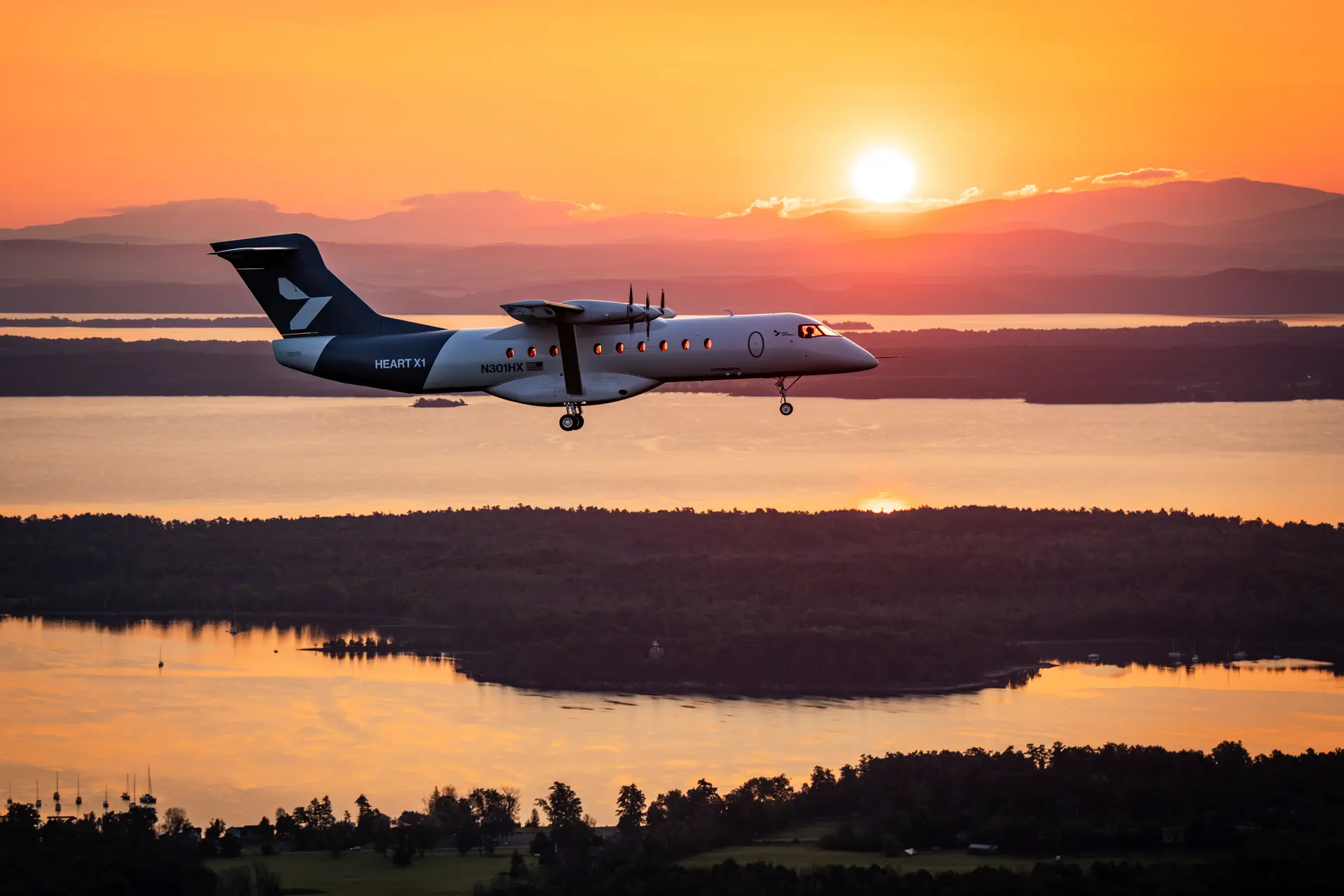 Twin-engine turboprop aircraft in landing/approach over a sunset-lit landscape with water and hills in the background.