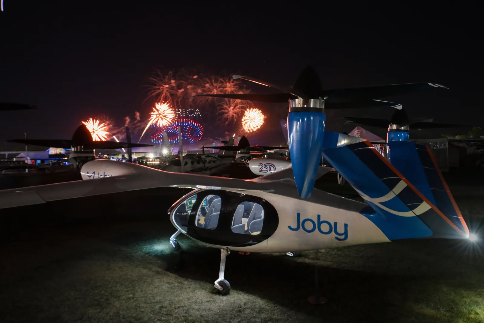 Night scene at an air show with a Joby eVTOL aircraft in the foreground and colorful fireworks in the background.
