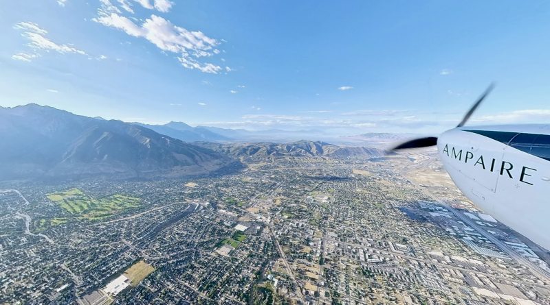 Aerial view from a small plane: city below with grid-like streets, distant mountains on the horizon, and part of the wing with 'AMPAIRE' visible on the right edge.